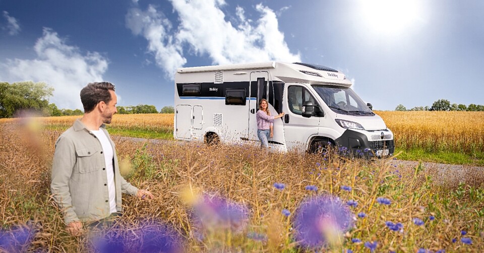 Weißes Hobby Wohnmobil auf einer sonnigen Straße im Kornfeld mit glücklichem Pärchen.